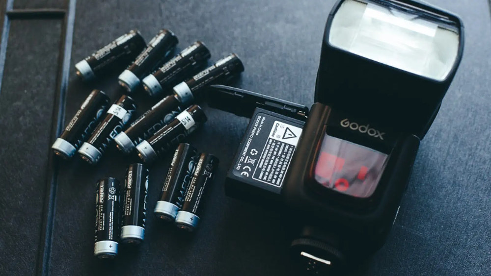 Вспышка Godox с открытым батарейным отсеком рядом с комплектом AA-аккумуляторов